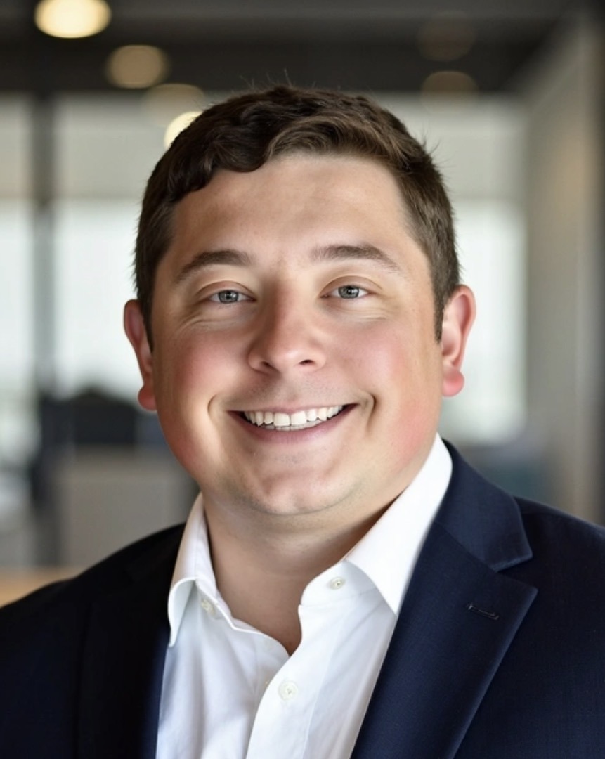 Smiling man in a suit in an office setting. He has short, dark hair and a white shirt. The tone is professional and approachable.