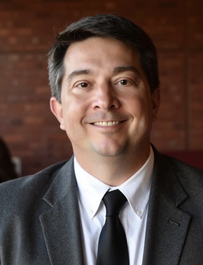 A man in a gray suit, white shirt, and black tie smiles warmly against a blurred brick wall background. The setting conveys a professional yet relaxed atmosphere.