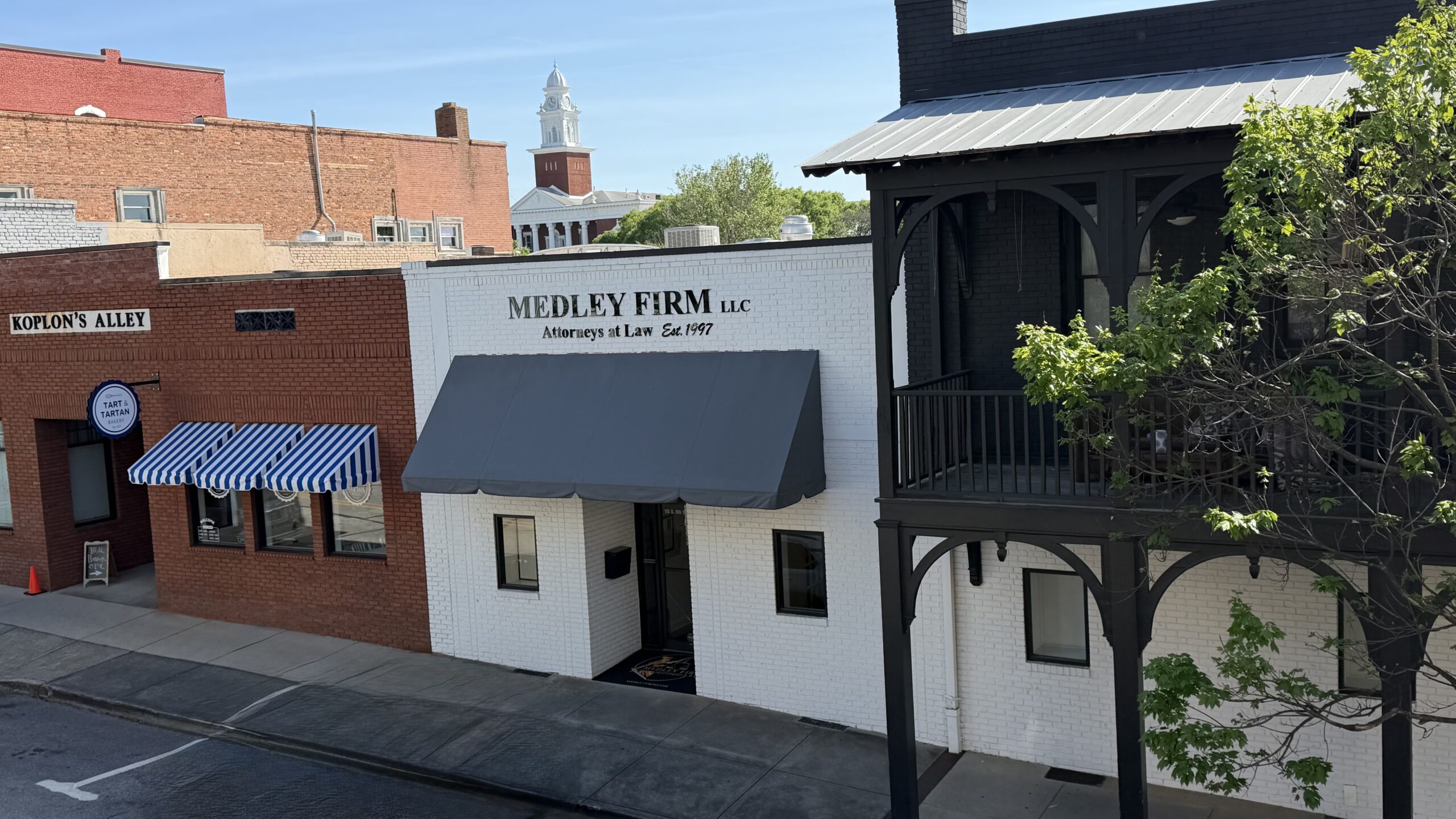 Street view of "Medley Firm LLC" in a white brick building with a dark awning. Adjacent is a red brick structure with a blue-striped awning. The scene is calm and sunny.