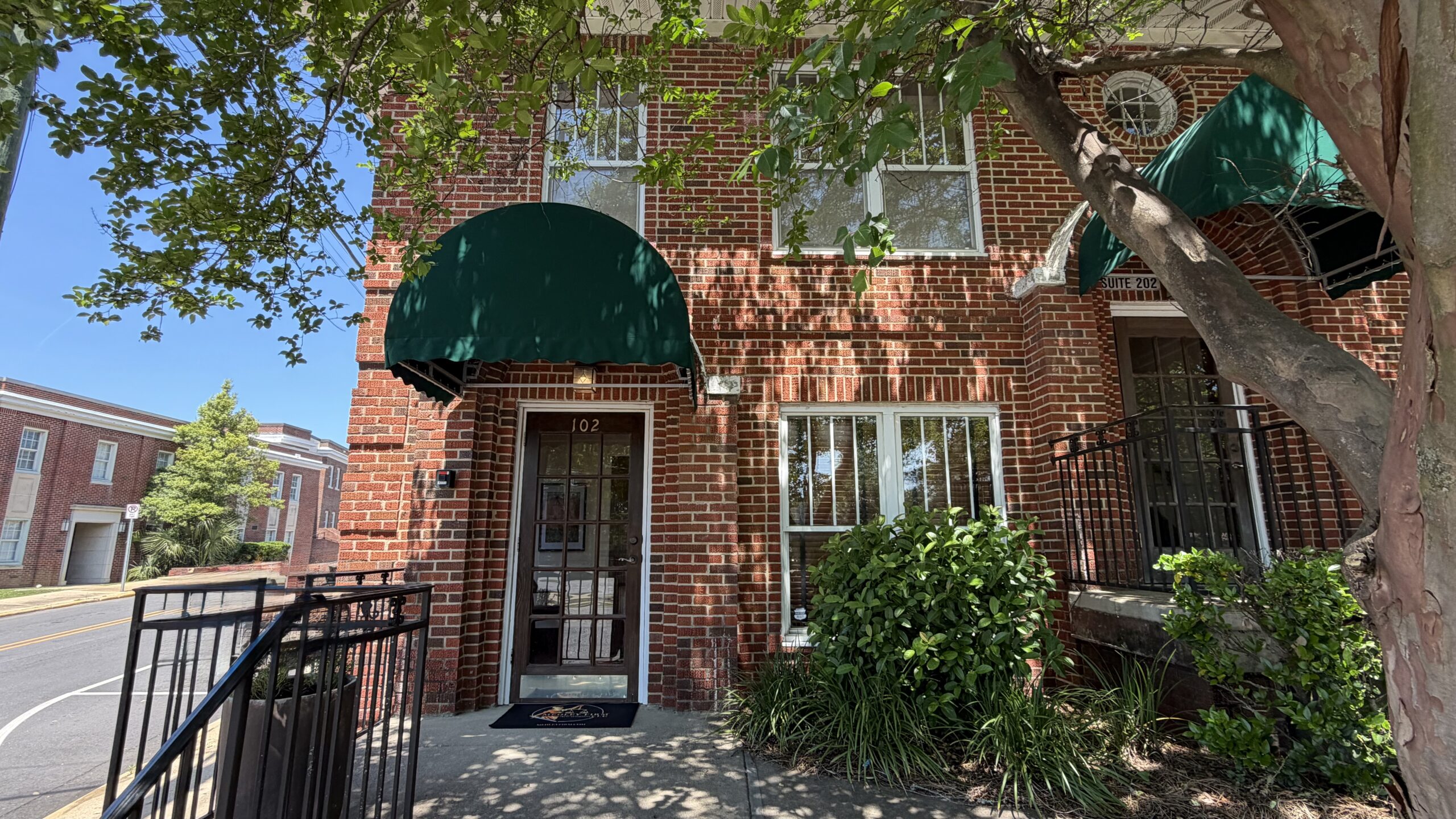 A brick building with green awnings shades the windows and door, set on a tree-lined street. Sunlight casts dappled shadows, creating a warm, inviting ambiance.